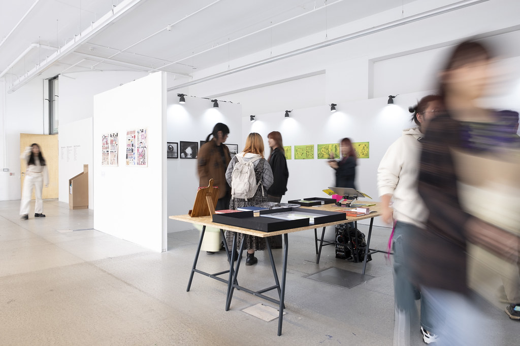 Visitors gathering around tables in a bright postgraduate exhibition